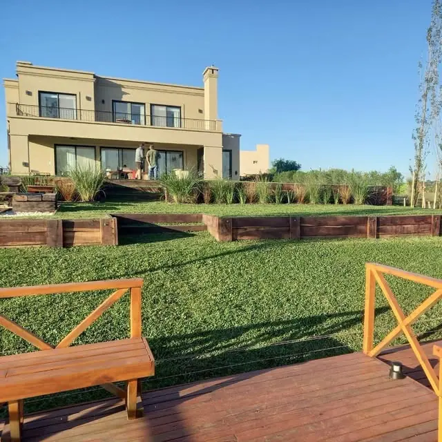Parque con deck de madera, bancos y taludes con durmientes frente a una casa
